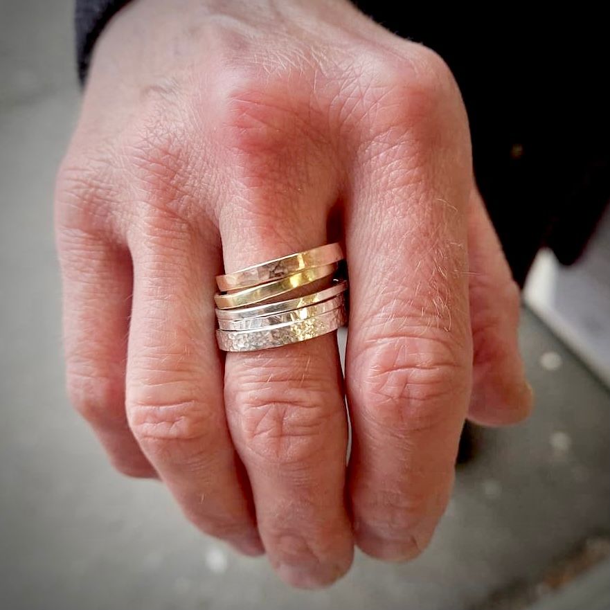A hand with 5 hammered rings stacked on middle finger. Rings are brass, copper & silver.