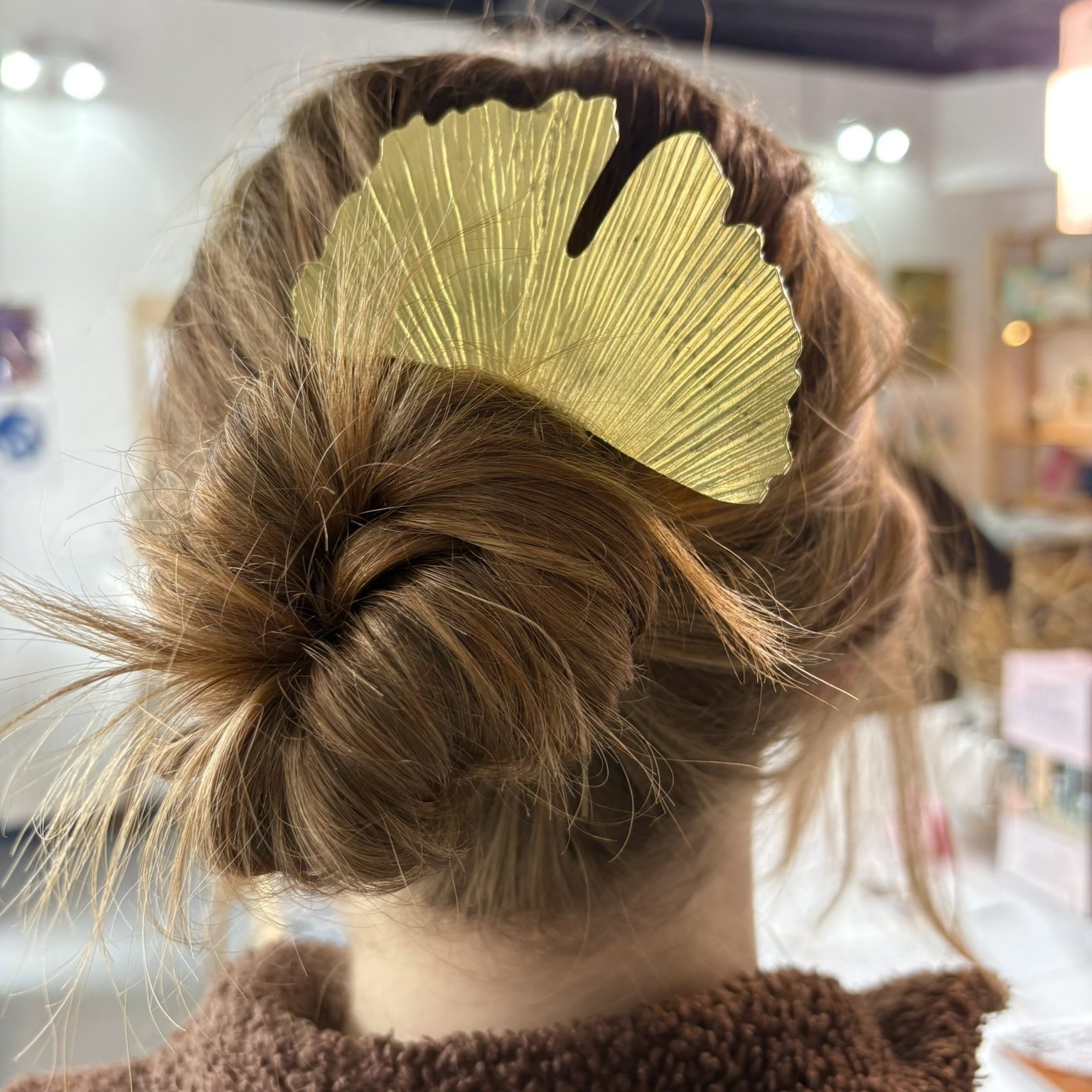 Image shows the back of a girls head, with her hair twisted into a bun and held in place with a large brass hair pin with a Gingko leaf design.