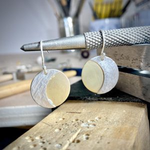 A pair of brass circle earrings with silver crescent moons, handing from a jewellery making tool